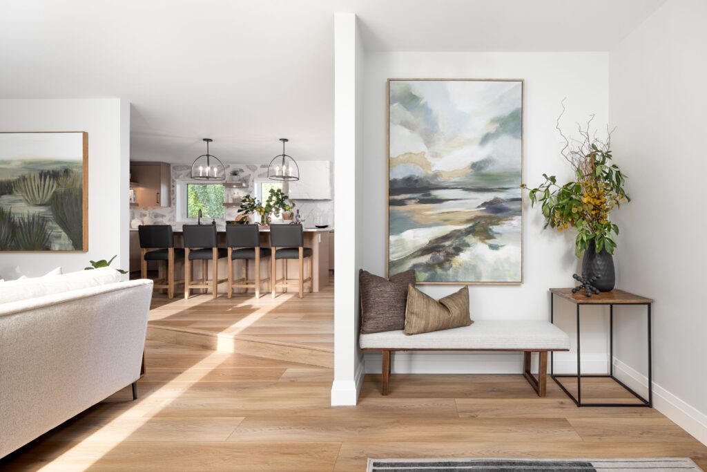 A modern, bright living space with light wood floors, a bench with pillows, a landscape painting, and a vase with flowers. The open kitchen and dining area with bar stools are visible in the background.