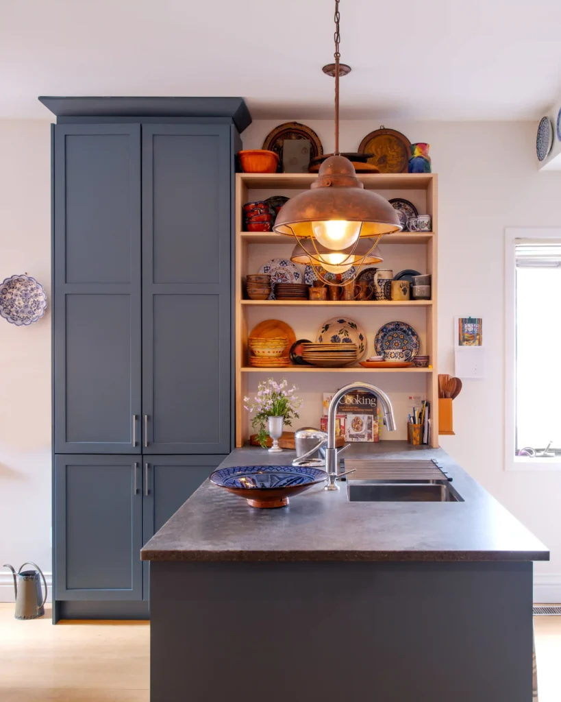 A modern kitchen with a gray island and sink, tall blue cabinets, open shelves filled with colorful dishes and pottery, and a large pendant light hanging above the counter.
