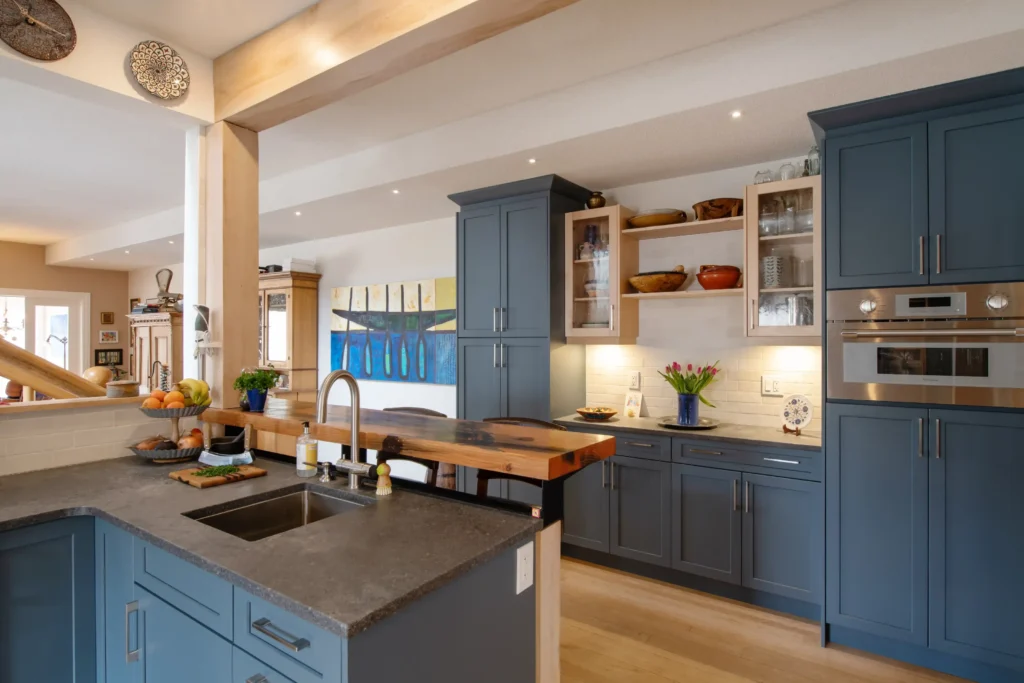 Modern kitchen with blue cabinets, wood countertops, and stainless steel appliances. Open shelves display bowls and plants; a sink sits on a dark countertop. Art and flowers add color to the bright, open space.