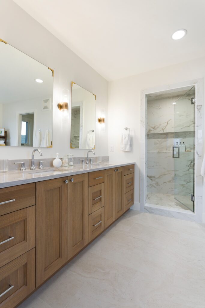 Modern bathroom with double sinks, wooden cabinets, two mirrors, wall sconces, and a glass-enclosed marble shower. The space is bright with neutral tones and minimalistic decor.