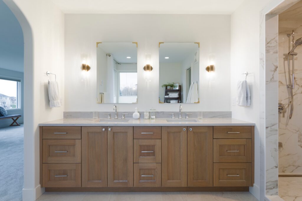 A modern bathroom with a double-sink wooden vanity, two mirrors, wall sconces, and towels on either side. A glass shower is visible on the right, and a bedroom can be seen through an archway on the left.