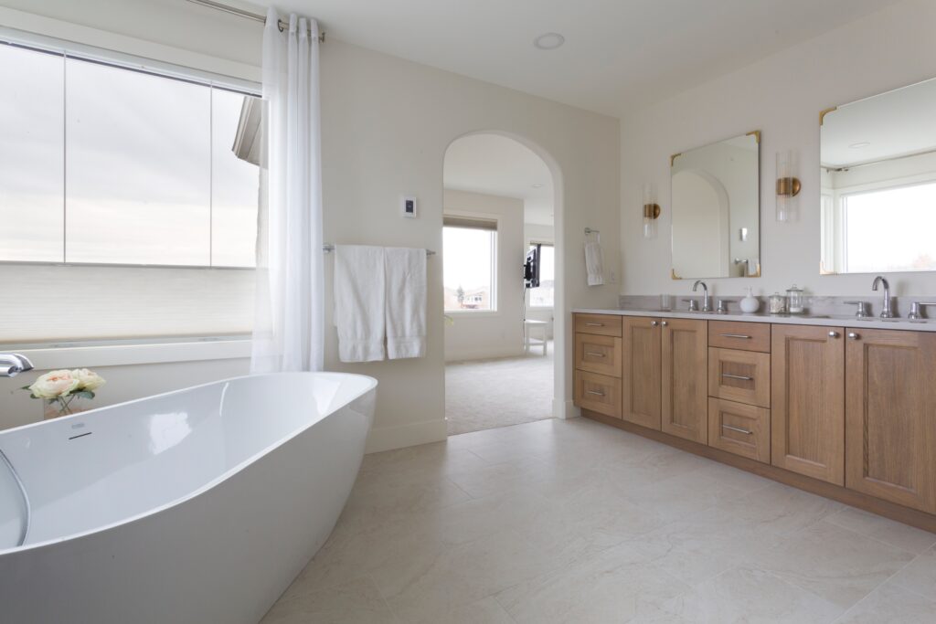 Modern bathroom with a freestanding white tub, wooden double-sink vanity, two mirrors, large windows, and neutral-colored walls and floor. Bright natural light fills the space, creating an open and airy feel.