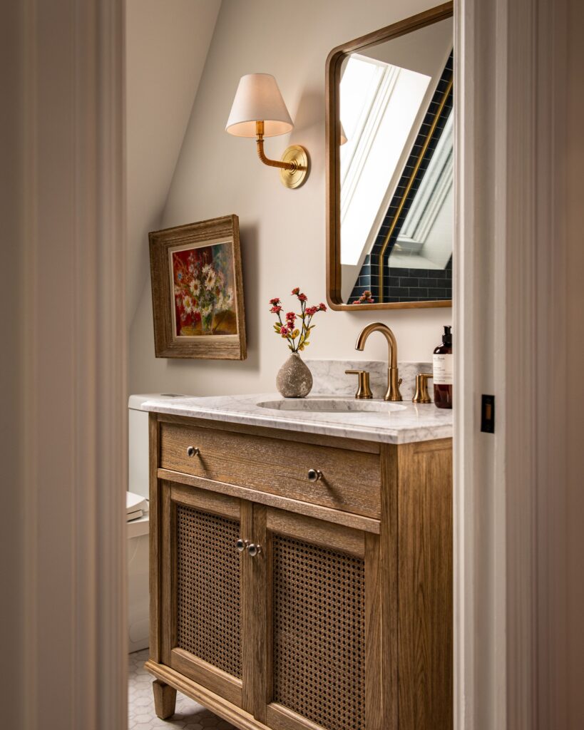 A warm, elegant bathroom with a wooden vanity, marble countertop, brass faucet, wall-mounted mirror, sconce light, small vase of red flowers, and a framed floral painting on the wall. Skylight reflection appears in the mirror.