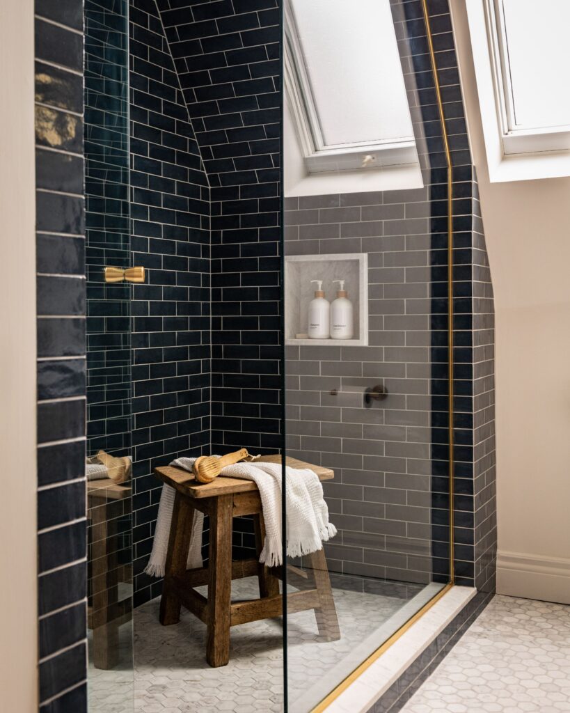 A modern shower with dark subway tiles, a glass door, and gold trim. A wooden stool with a towel and brush sits inside. A built-in shelf holds white bottles. Light enters through a slanted skylight above.