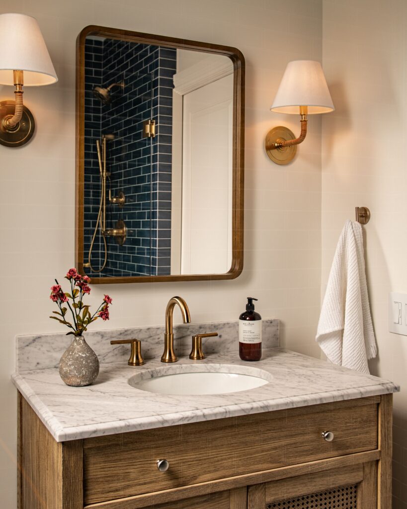 A bathroom vanity with a marble countertop, gold faucet, and wooden cabinet. A rectangular mirror is flanked by two wall sconces. A towel hangs on a hook, and a vase with flowers and soap bottle sit on the counter. Blue tiled shower reflected.