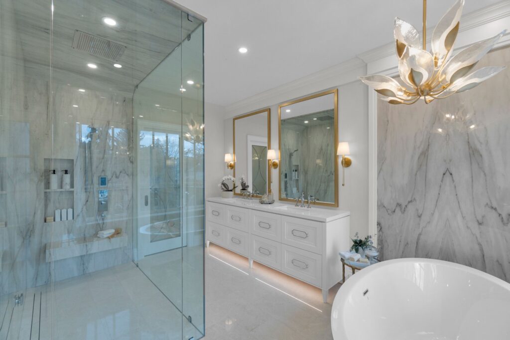 Luxurious modern bathroom with a glass-enclosed shower, double vanity with gold-framed mirrors, a freestanding bathtub, marble walls, and a sculptural chandelier. The space is bright and elegant.