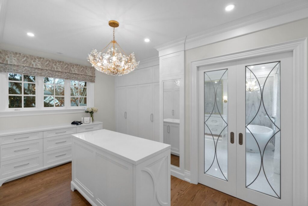 A luxurious white walk-in closet with an island, built-in cabinets, a crystal chandelier, hardwood floors, a window with patterned valance, and glass French doors revealing a bathroom with a freestanding tub.