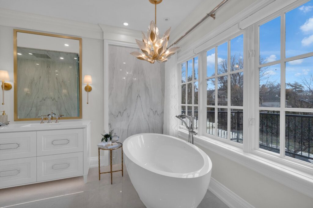 Bright, elegant bathroom with a white freestanding tub, large window, marble accents, a gold-framed mirror, wall sconces, and a decorative modern chandelier. Natural light fills the room.