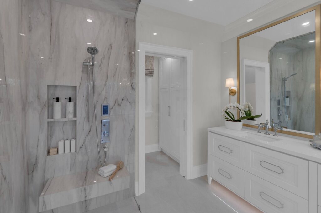 Modern bathroom with a marble shower featuring a built-in bench and shelves, a white vanity with a large gold-framed mirror, a wall sconce, an orchid plant, and sleek minimalist decor.