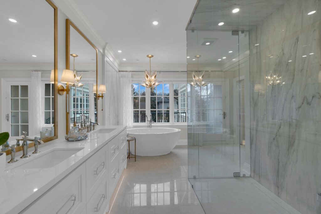 A modern, luxurious bathroom featuring a large white bathtub by a window, glass-enclosed shower, marble walls, double sinks with mirrors, and elegant lighting fixtures.