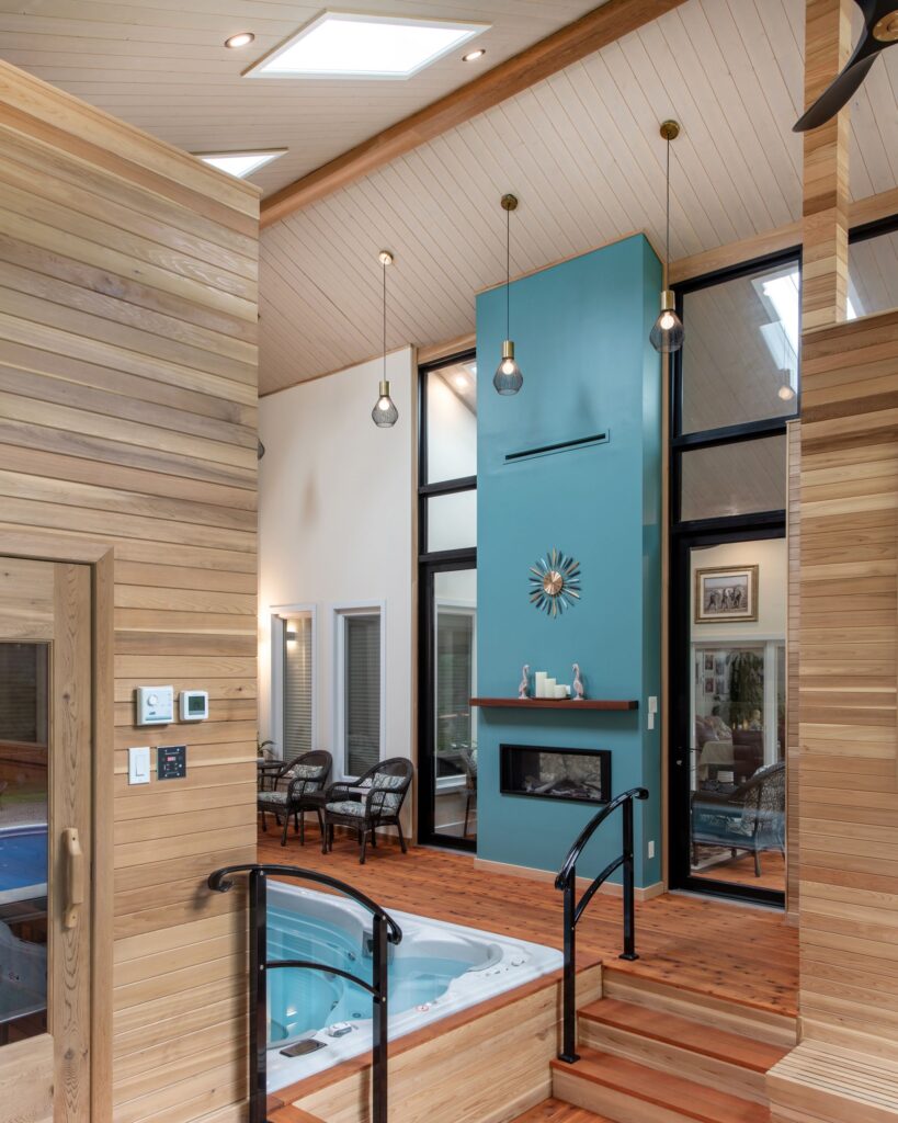 A modern indoor space features a sunken hot tub, wood-paneled walls and ceiling, pendant lights, a blue accent wall with a fireplace, and a seating area with wicker chairs and large windows.