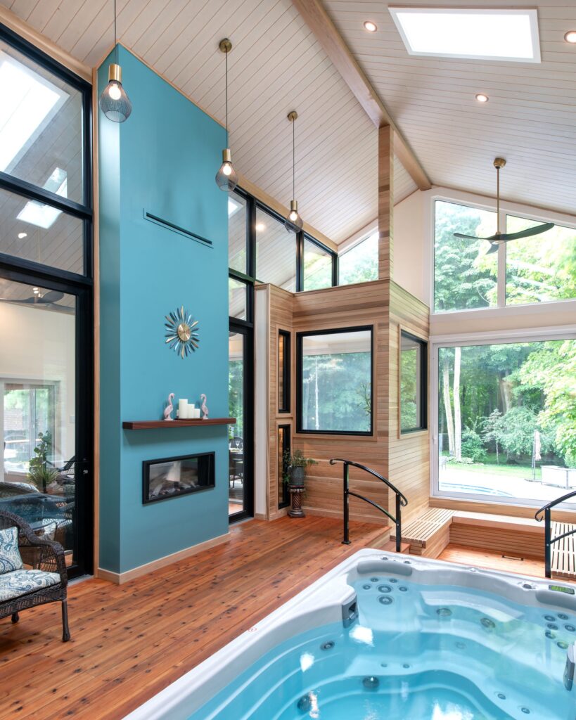 Modern indoor sunroom with a hot tub, high angled ceilings, large windows, light wood walls and floor, a blue accent wall with a fireplace and decor, and views of a green backyard.