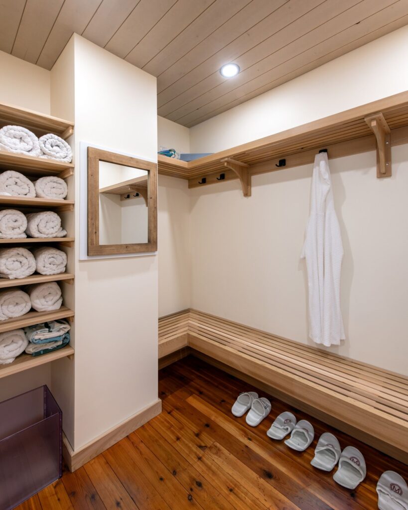 A tidy locker room with wooden benches, shelves stacked with rolled white towels, a hanging white robe, a wall mirror, and four pairs of white slippers neatly arranged on polished wooden floors.