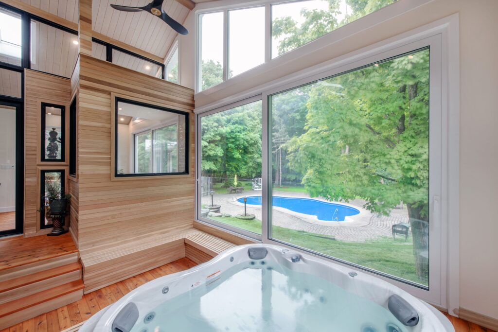 Modern indoor spa room with a hot tub next to large windows overlooking a lush green backyard and an outdoor swimming pool surrounded by trees. Natural light fills the space with wooden accents.