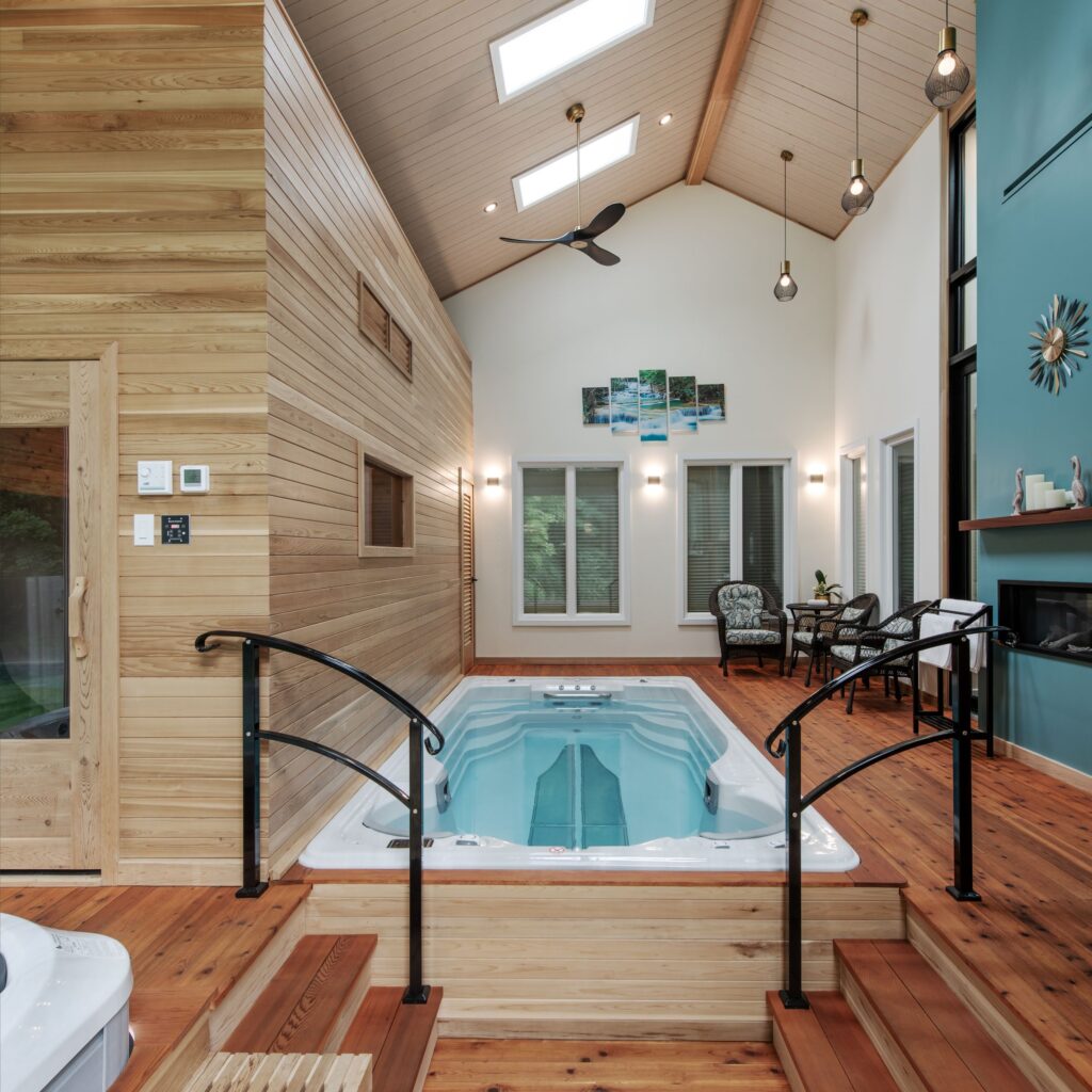 A modern indoor pool room with wood-paneled walls and ceiling, large windows, skylights, a sauna, and a seating area with chairs and a table beside a turquoise accent wall and fireplace.