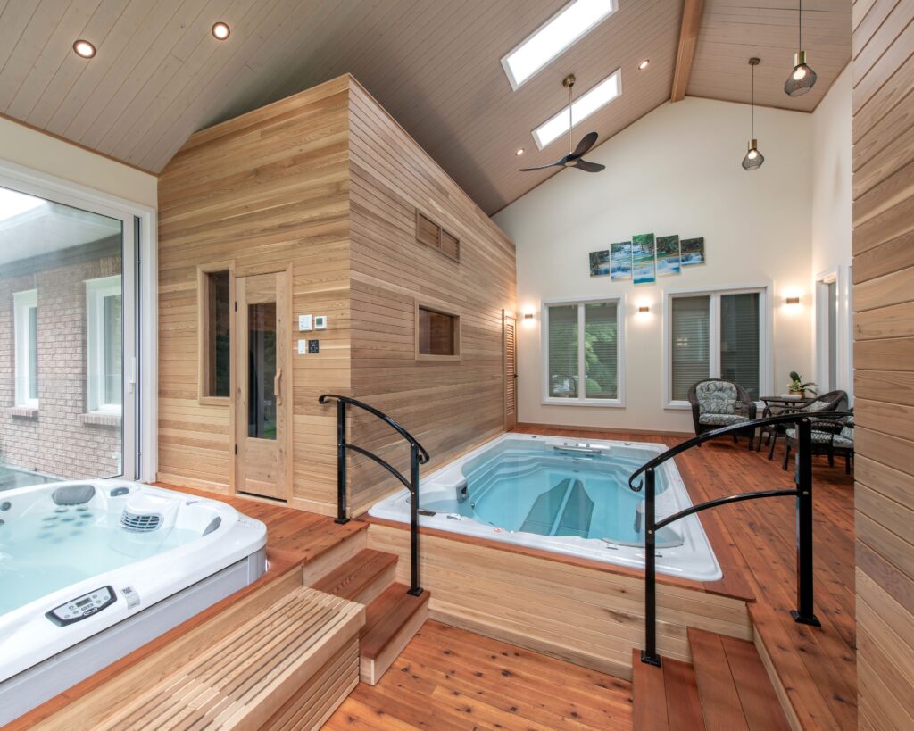 A modern indoor spa room with a hot tub and pool, wood-paneled walls and floor, skylights, large windows, and a small seating area with chairs and a table.