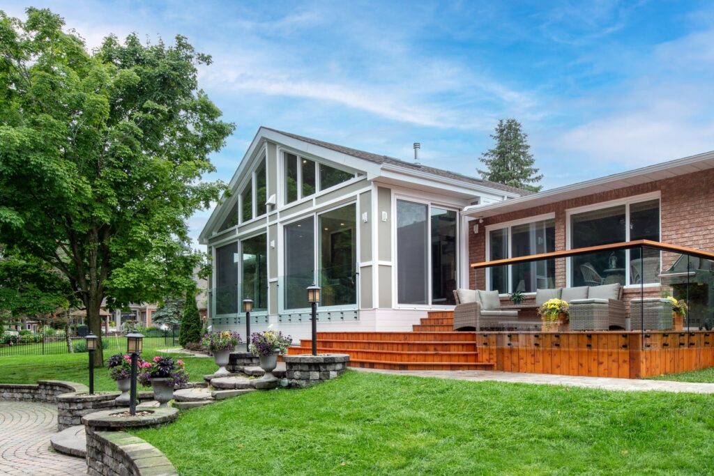 A modern house with large windows, a sunroom, and an elevated wooden patio with outdoor seating, surrounded by green grass, trees, and landscaped stone steps on a sunny day.