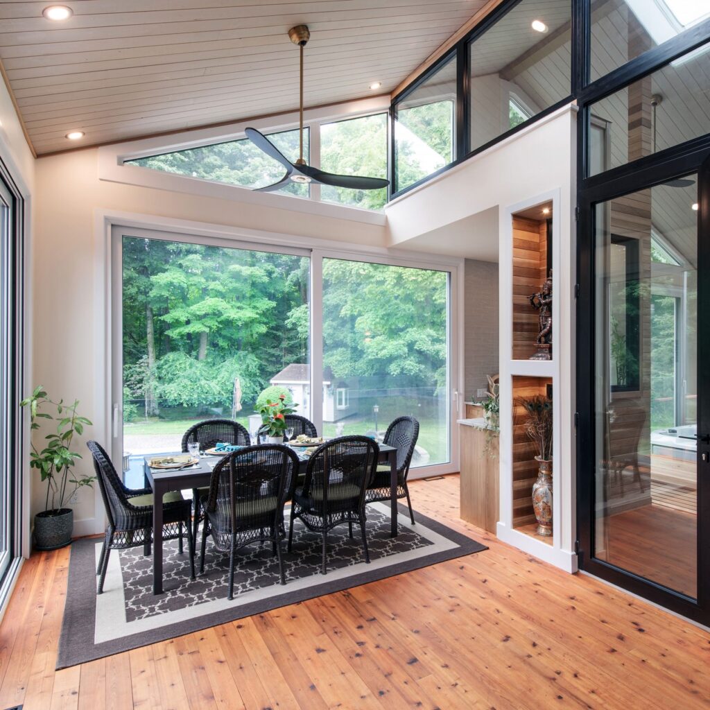 A bright sunroom with large windows, wooden floors, and a black dining set for six. Green trees and a yard are visible outside. The room features a ceiling fan and indoor plants, creating a cozy, modern atmosphere.