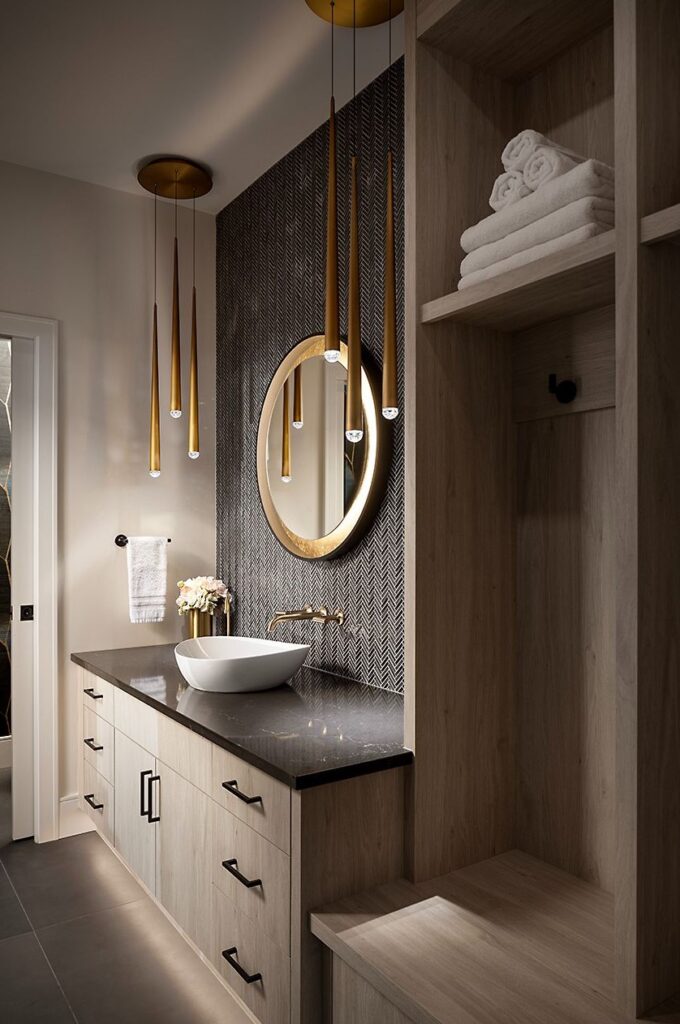 Modern bathroom with wood cabinetry, a round mirror above a vessel sink, pendant lights with gold accents, folded white towels on shelves, and a textured dark wall behind the sink.