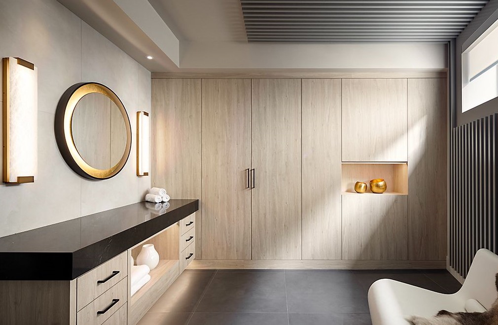 A modern bathroom with light wood cabinets, a black countertop, a round mirror with gold trim, soft lighting, open shelving with towels, built-in shelves holding gold vases, and large gray floor tiles.