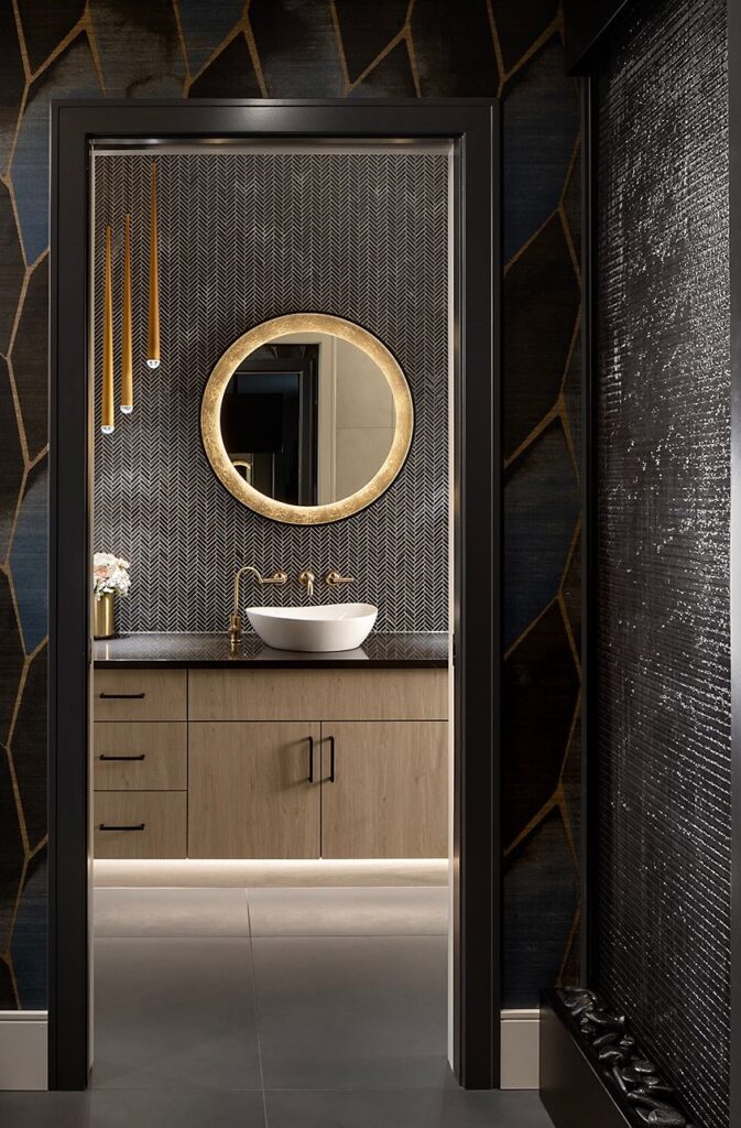A modern bathroom features a round, illuminated mirror above a white vessel sink on a wooden vanity with gold fixtures, textured dark wallpaper, pendant lights, and a nearby water feature.
