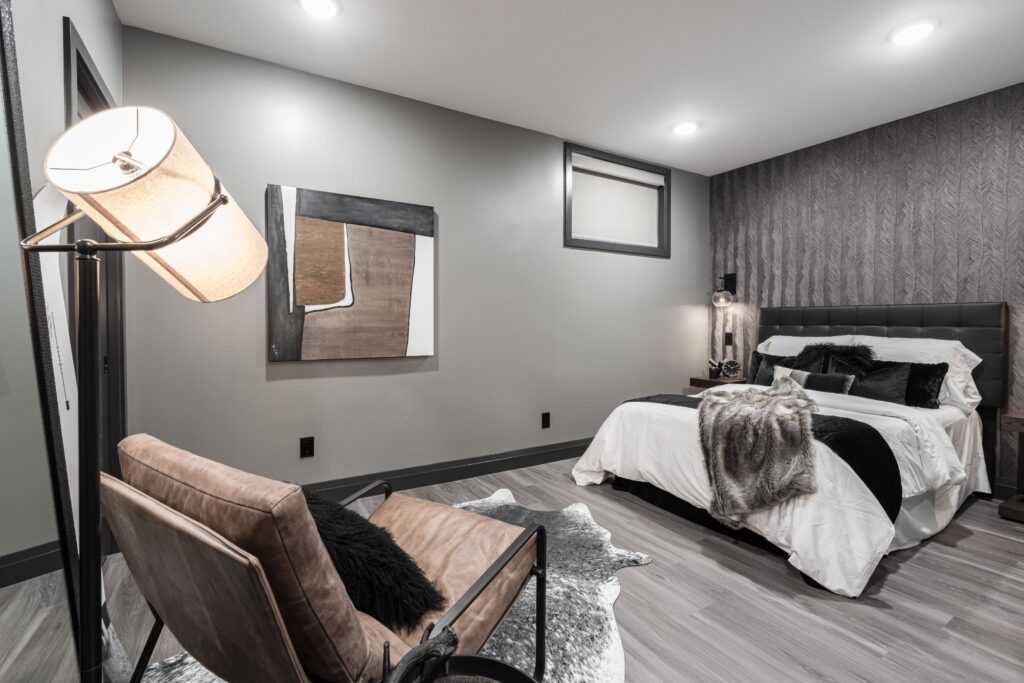 Modern bedroom with a neutral color palette, featuring a bed with white bedding and dark accents, a brown accent chair with a floor lamp, contemporary artwork on the wall, and a cozy, minimalist decor.