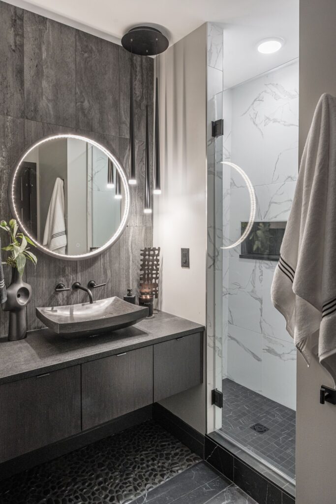 A modern bathroom with a round, backlit mirror above a dark stone sink and countertop, black fixtures, and a glass shower with marble-patterned walls. A striped towel hangs on the wall.