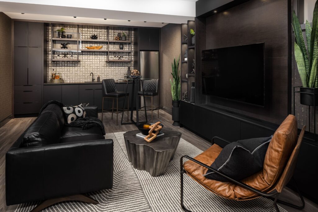 Modern open-concept living room and kitchen with dark cabinetry, a black leather sofa, brown accent chair, geometric coffee table, wall-mounted TV, bar seating, and decorative plants against a stylish, neutral background.