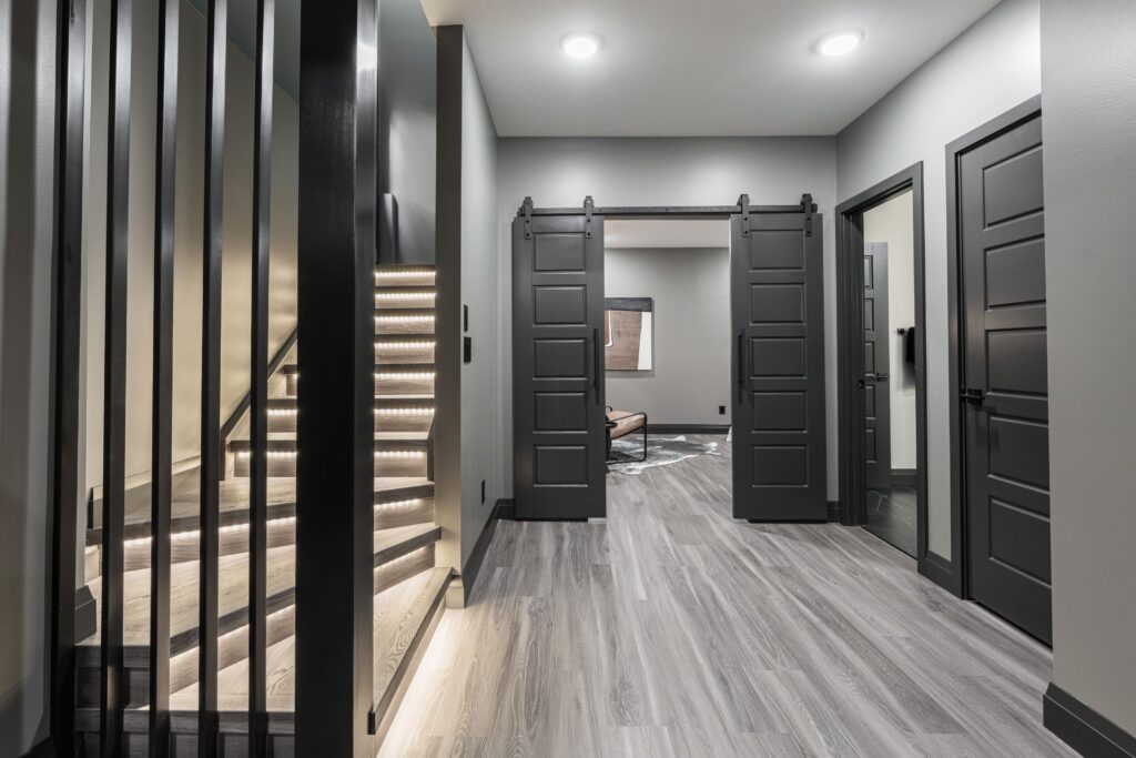 Modern hallway with gray walls and flooring, featuring a staircase with illuminated steps on the left, and three black doors, including double sliding barn doors opening to a room with a window and a chair.