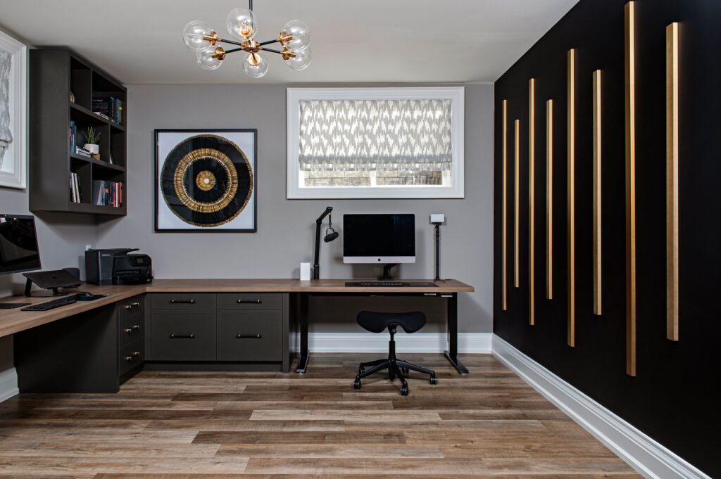 Modern home office with an L-shaped wooden desk, computer, black stool, wall shelves, and a decorative black wall with vertical gold accents. A round artwork and a window with patterned curtains are featured.
