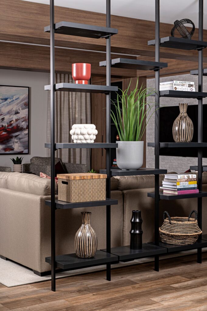 A modern black open shelving unit with decorative vases, a red bowl, a basket, a gray planter with green leaves, and other decor items separates a living room with taupe sofas and a colorful abstract painting.