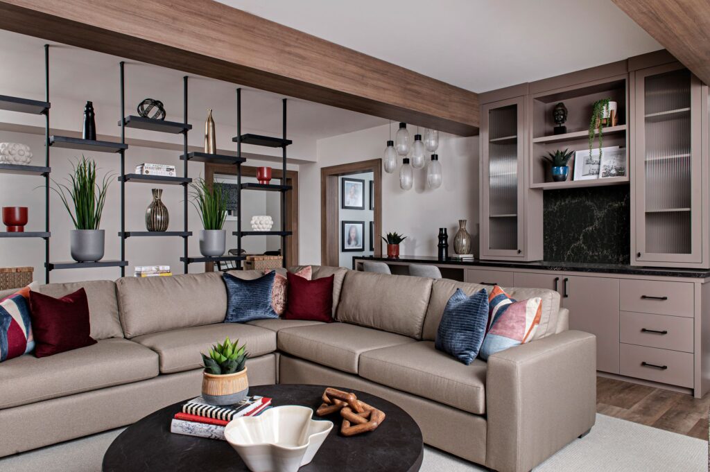 A modern living room with a beige sectional sofa decorated with colorful pillows, a round coffee table, open shelving with plants and decor, built-in cabinets, and pendant lights. The space is bright and stylish.