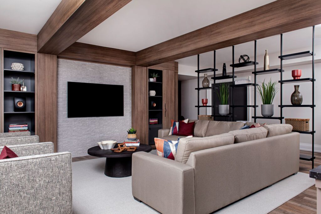 Modern living room with a large beige sectional sofa, round black coffee table, built-in shelving with decor, wall-mounted TV, and neutral tones accented by pillows and greenery.