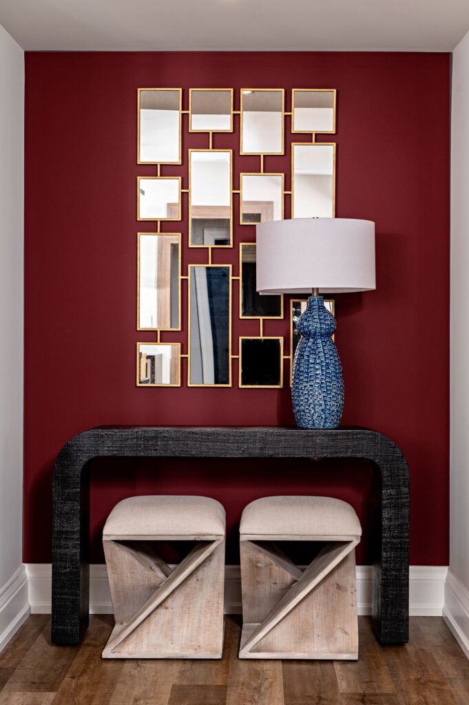 A modern entryway features a geometric black console table, two beige cube stools, a blue patterned table lamp, and a wall-mounted grid of rectangular mirrors with gold frames against a deep red wall.
