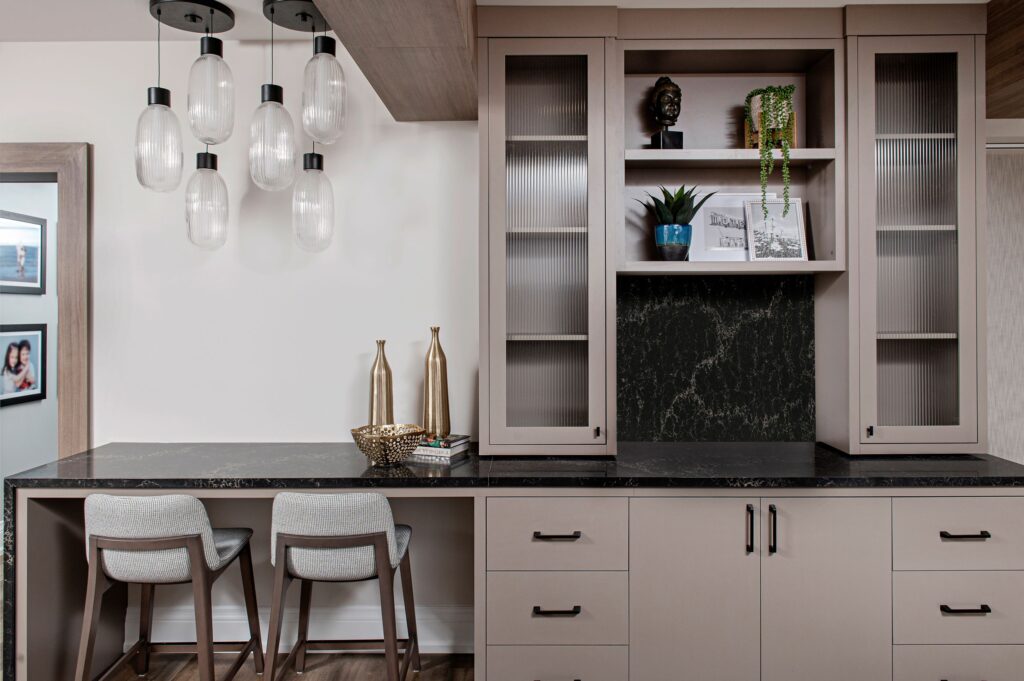 Modern home interior with a black countertop, beige cabinets, two gray chairs, hanging glass pendant lights, decorative vases, plants, photos, and a small statue on open shelves.