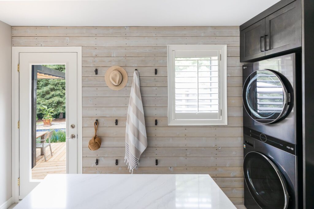 A modern laundry room with a white wood paneled wall, a window with shutters, a stacked black washer and dryer, a striped towel, a straw hat, and a brush hanging on hooks, and a door leading outside.