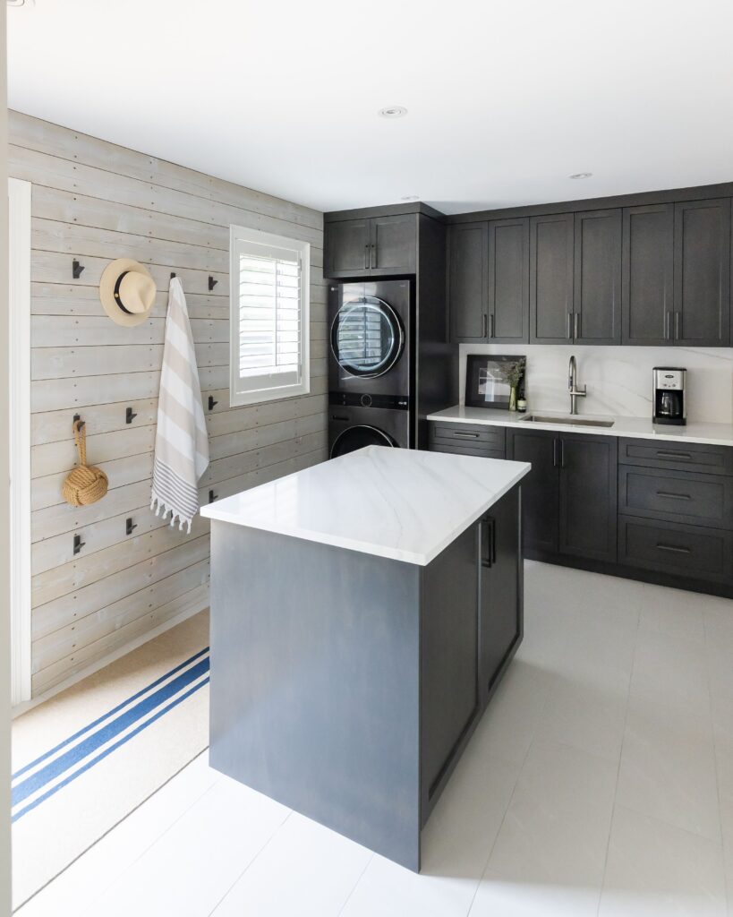 A modern laundry room with dark cabinets, a marble-topped island, stacked washer and dryer, and white plank walls. Hooks hold a towel, hat, and brush. A window lets in natural light.
