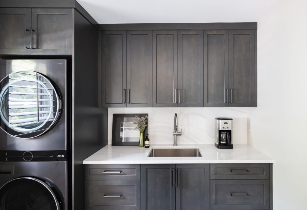 Modern laundry room with dark gray cabinets, a stacked washer and dryer on the left, a countertop with a sink, and a small framed picture with a vase of flowers placed near the backsplash.