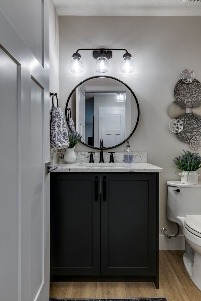 A modern bathroom features a black vanity with a marble countertop, a round mirror, and three-bulb light fixture above. Decorative towels, plants, and wall art add accents. A toilet is visible beside the vanity.