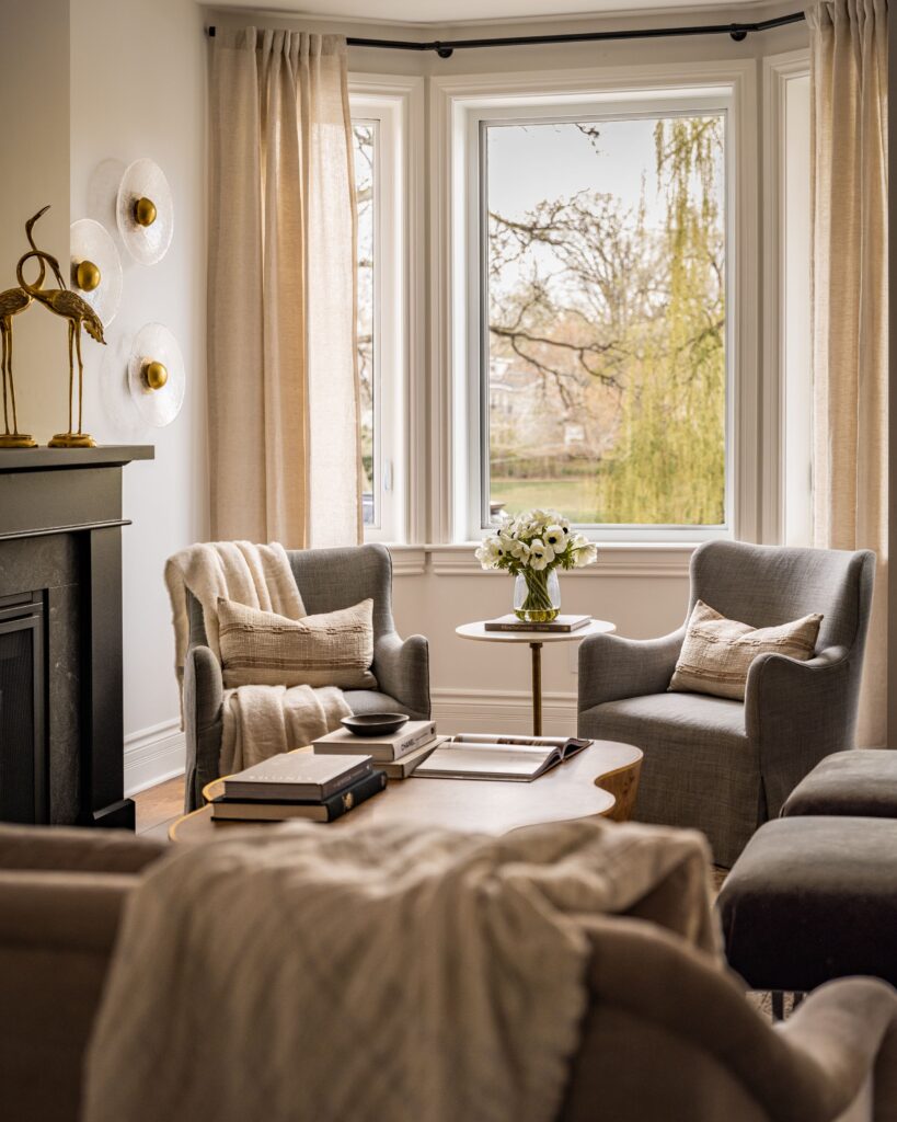 A cozy living room with two gray armchairs by a bay window, beige curtains, a small round table with a flower vase, neutral-toned decor, and soft natural light filling the space.
