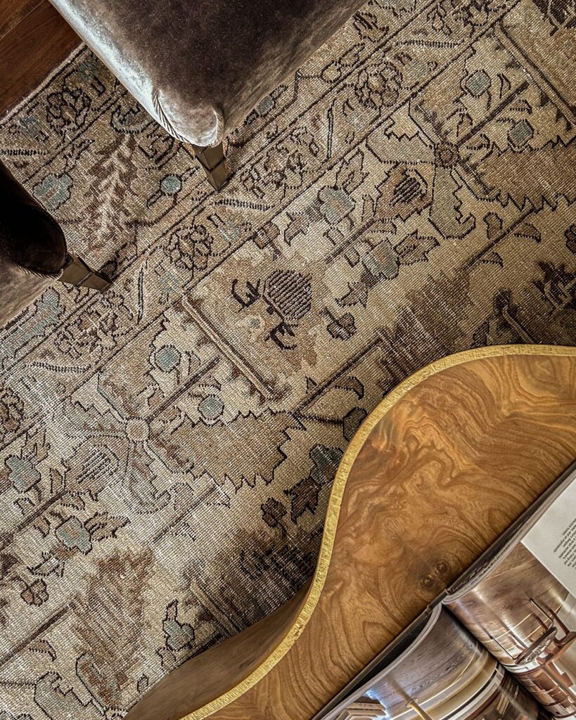 A close-up view of a patterned, vintage-style rug with brown tones, partially covered by the corner of a velvet chair and the curved edge of a wooden table with an open book on it.