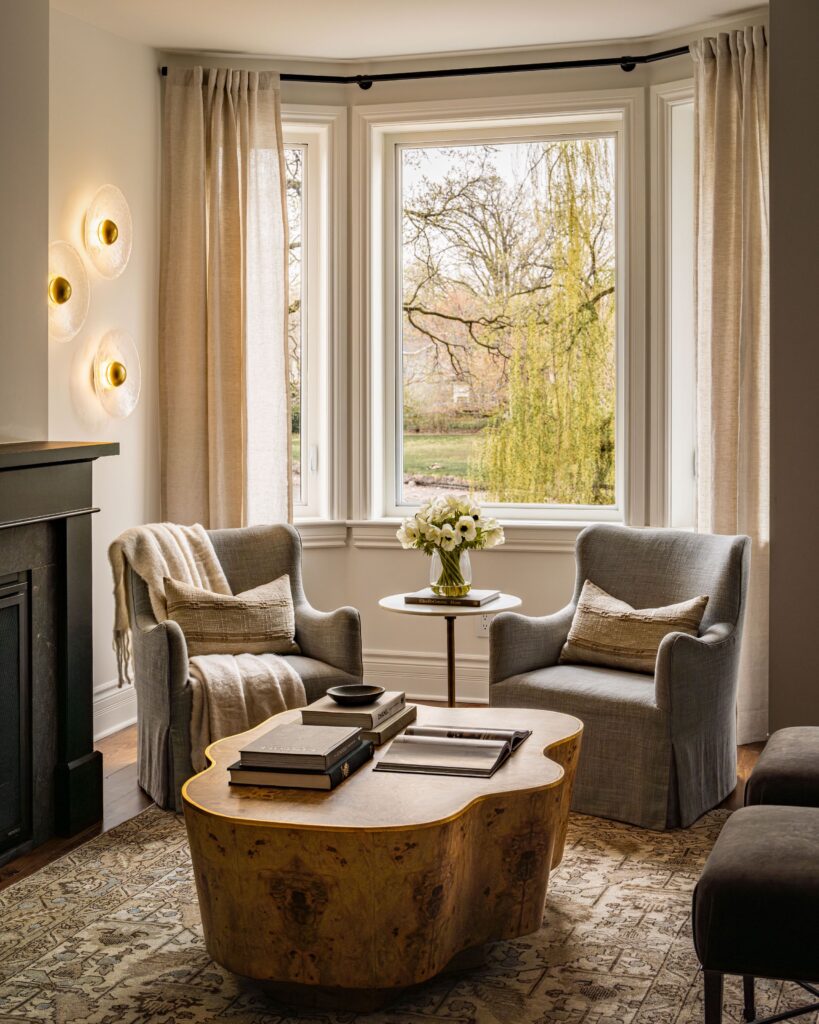 A cozy sitting area with two gray armchairs, a live-edge wooden coffee table, books, and a vase of flowers by a large bay window with beige curtains. Soft wall lights and a leafy outside view create a warm atmosphere.