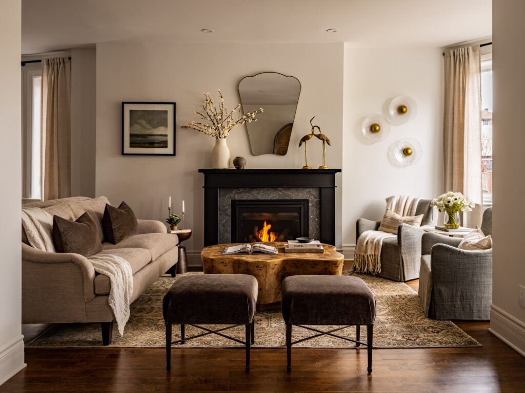 A cozy living room with a fireplace, beige sofa, two gray armchairs, a wooden coffee table, two dark ottomans, and decorative items. Warm lighting, neutral tones, and a large mirror above the fireplace.