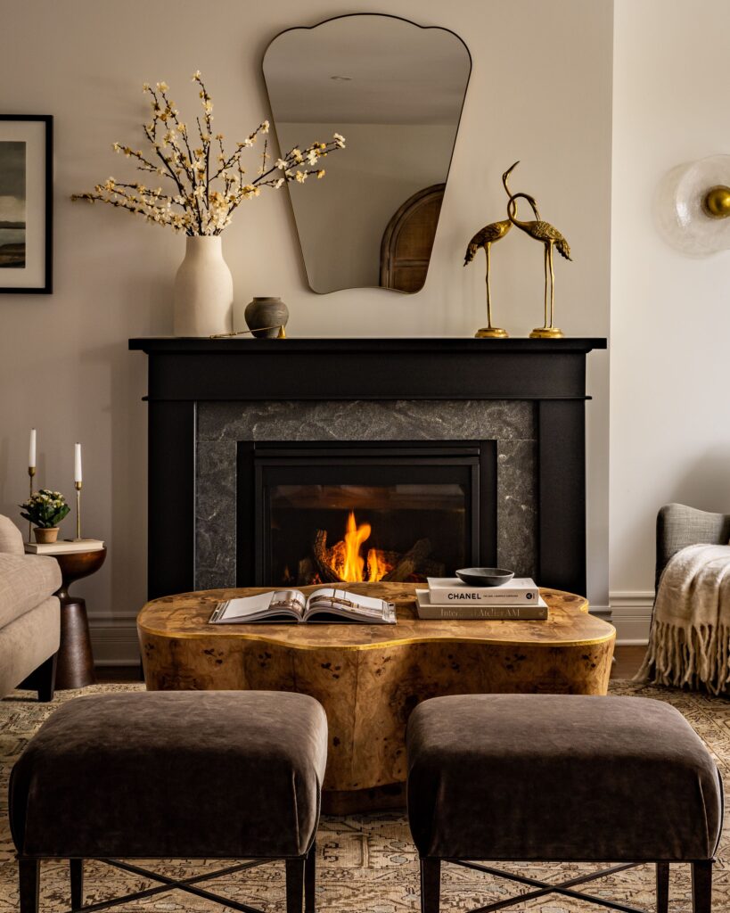 A cozy living room with a lit fireplace, a curved wooden coffee table holding books, two brown ottomans, and decorative items on the mantel including a vase with flowers, gold crane sculptures, and a mirror above the mantel.