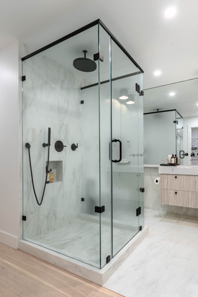 A modern bathroom featuring a glass-enclosed shower with black fixtures, white marble walls and floor, a built-in shower niche, and a rain showerhead. A vanity with a large mirror is visible in the background.
