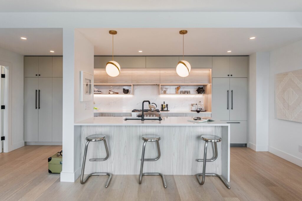 A modern kitchen with a white island, three metal stools, two round pendant lights, built-in cabinets, open shelves, and light wood flooring. A suitcase is on the floor near the left doorway.