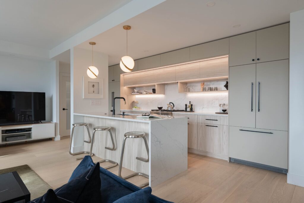 Modern kitchen with light wood cabinets, a marble island with three metal bar stools, built-in appliances, globe pendant lights, open shelving, and a living area with a TV and blue sofa in the foreground.
