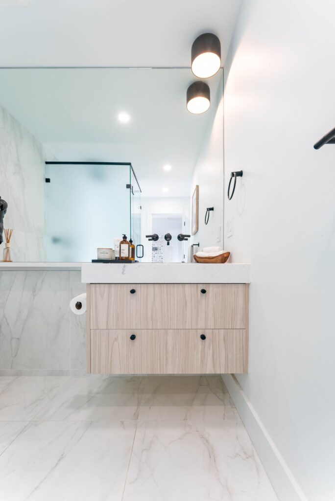 Modern bathroom with light wood floating vanity, wall-mounted faucet, large mirror, marble floor and walls, glass shower enclosure, and two black cylindrical wall lights above the mirror. Minimalist and bright design.