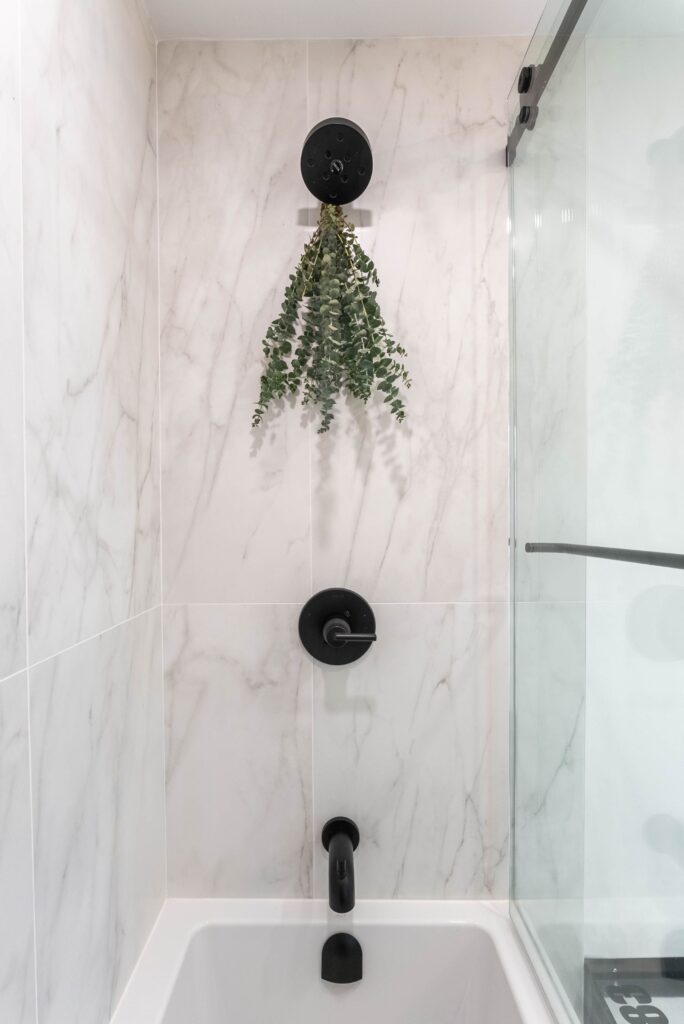 A modern bathtub with white marble walls, black fixtures, and a glass shower door. A bundle of green eucalyptus hangs from the showerhead above the tub.