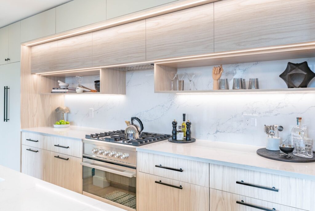 A modern kitchen featuring light wood cabinets, a gas stove with a kettle, marble backsplash, open shelves with decor items, and neatly arranged kitchen accessories on a white countertop.